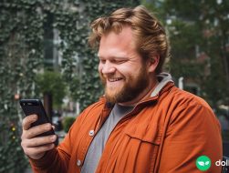 A large man looking down at a cell phone in his hand with a smile on his face. Greenery in the background.