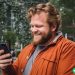 A large man looking down at a cell phone in his hand with a smile on his face. Greenery in the background.