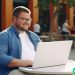 A slightly overweight man sitting at a small table outside with a laptop. He is smiling.