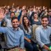 A large group of employees at a new employee orientation. They are all raising their hands.