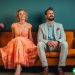 A classy married couple sitting on a couch but slightly facing away from each other. They have playful looks on their faces.
