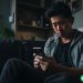 A young Asian man sitting on his couch, comparing two apps on his phone.