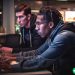 A young tech worker fervently typing on his keyboard while his coworker sits by anxiously watching and hoping he can fix the problem.